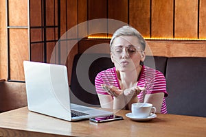 Freelancer with blonde short hair, in pink t-shirt and eyeglasses is sitting alone in cafe and working on laptop and sending air