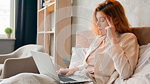 Freelancer Caucasian woman working remote distant from home in bed thinking pensive holding head girl with laptop work