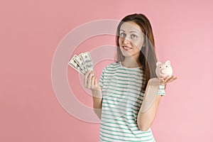 A freelancer girl in casual clothes smiles and holds hundred dollar bills and a piggy bank in her hands standing on a pink