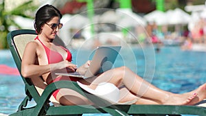 Freelancer girl in red swimsuit working on laptop pc lying on deck chair near swimming pool