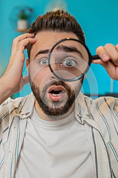 Freelancer man holds magnifying glass near face looking at camera with big zoomed eye, analyzing