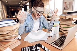 Freelancer man looking at notes frustrated at laptop sitting at desk surrounded by books.