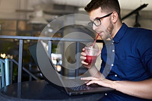Freelancer man with plastic cup of fresh beverage in hand works with laptop. Businessman in glasses drinks juice for