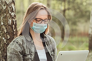 Freelancer during quarantine in the nature. Woman in mask is working on laptop in forest.