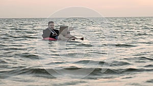 Freelancer swims in the open sea on a children`s inflatable circle and works with a laptop on his knees.