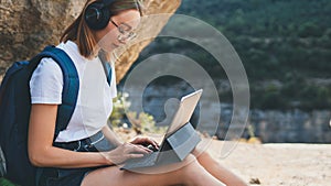 Freelancer woman with glasses and headphones uses digital tablet device working in nature outdoor, tourist girl with backpack