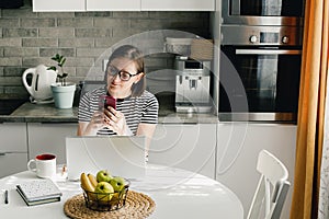 Freelancer woman wearing glasses using phone and laptop while working from home
