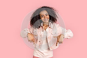 Freestyle. African girl in denim jacket standing isolated on pink pointing down at copy space smiling joyful