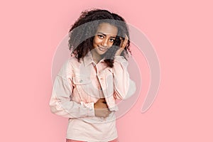 Freestyle. African girl in denim jacket standing isolated on pink posing touching hair smiling cute