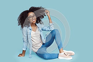Freestyle. African girl in glasses sitting isolated on gray holding curl posing smiling cheerful