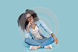 Freestyle. African girl in glasses sitting isolated on gray leaning on ahnd smiling relaxed