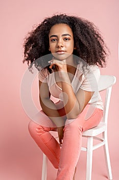 Freestyle. African girl sitting on chair isolated on pink leaning on hand smiling pensive close-up
