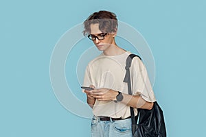 Freestyle. Man in glasses with bag standing isolated on gray browsing interent on smartphone concentrated