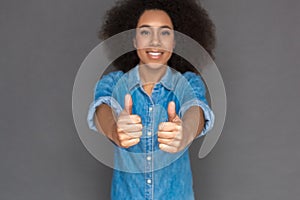 Freestyle. Mulatto woman standing on grey showing thumbs up close-up blurred smiling toothy