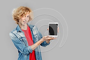 Freestyle. Teen blond boy standing isolated on gray showing screen on digital tablet copy space posing to camera playful