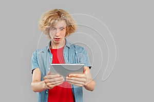 Freestyle. Teen blond boy standing isolated on gray watching video on digital tablet shocked