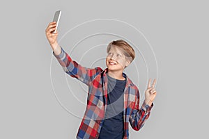 Freestyle. Young boy standing isolated on grey taking selfie posing to camera showing salute gesture happy