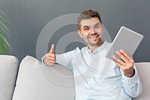 Freestyle. Young man studio sitting on sofa isolated on grey with digital tablet showing thumb up smiling cheerful