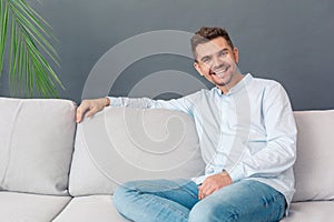 Freestyle. Young man studio sitting on sofa relaxed isolated on grey smiling joyful