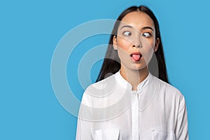 Freestyle. Young woman wearing shirt standing isolated on blue looking at nose showing tongue grimacing silly close-up