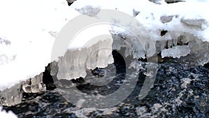 Freeze snowy big boulder in clear cold water of mountain river. Small icicles glitter above crystal water of noisy stream.