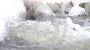 Freeze snowy big boulder in clear cold water of mountain river. Small icicles glitter above crystal water of noisy stream.