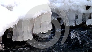 Freeze snowy big boulder in clear cold water of mountain river. Small icicles glitter above crystal water of noisy stream.