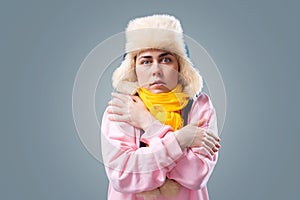 Freezing young caucasian woman wearing ushanka hat and scarf is hugging herself trying to keep warm. Gray background