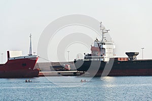 Freight truck stands between largo cargo ships in shipping harbor, oarsmen rowing in foreground sea, port Monopoli, Italy