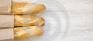 French baguettes in paper bags on white wooden background, top view. Space for text
