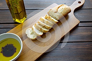 A french bread baguette sliced up on a cutting board with a bowl of olive oil with a splash of balsamic vinegar along side a bott