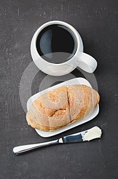 French breakfast. White mug with black coffee and croissant with butter knife on dark stone table. Top view. Coffee break