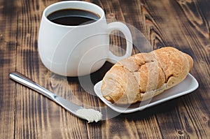 French breakfast. White mug with black coffee and croissant with butter knife on wooden table. Coffee break