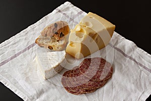 French brie and Swiss Emmental cheese with slices of salami sausage and a home baked bun placed on a white napkin background