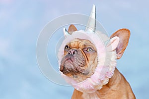 French Bulldog dog wearing funny unicorn costume headband in front of blue background