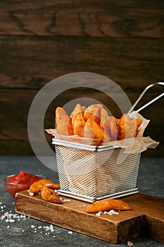 French fries in metal wire basket with salt and ketchup on old wooden dark background clous up. Fried potatoes. Fast food