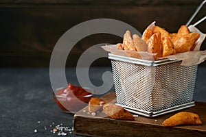 French fries in metal wire basket with salt and ketchup on old wooden dark background clous up. Fried potatoes. Fast food