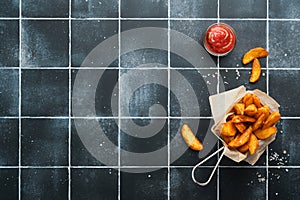 French fries in metal wire basket with salt and ketchup on old wooden dark background clous up. Fried potatoes. Fast food