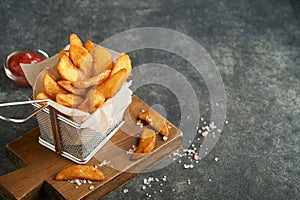 French fries in metal wire basket with salt and ketchup on old wooden dark background clous up. Fried potatoes. Fast food