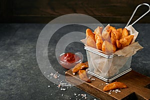 French fries in metal wire basket with salt and ketchup on old wooden dark background clous up. Fried potatoes. Fast food and