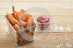 French fries in metal wire basket with salt and ketchup on old wooden light background clous up. Fried potatoes. Fast food