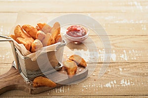 French fries in metal wire basket with salt and ketchup on old wooden light background clous up. Fried potatoes. Fast food