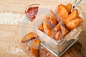 French fries in metal wire basket with salt and ketchup on old wooden light background clous up. Fried potatoes. Fast food