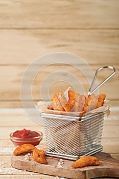 French fries in metal wire basket with salt and ketchup on old wooden light background clous up. Fried potatoes. Fast food