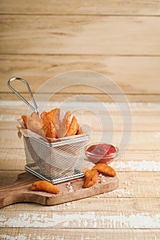 French fries in metal wire basket with salt and ketchup on old wooden light background clous up. Fried potatoes. Fast food