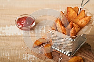 French fries in metal wire basket with salt and ketchup on old wooden light background clous up. Fried potatoes. Fast food