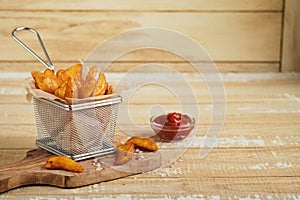 French fries in metal wire basket with salt and ketchup on old wooden light background clous up. Fried potatoes. Fast food