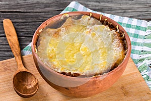 French onion gratin soup in a clay pot, authentic recipe, wooden spoon on a cutting board on an old rustic table, close-up