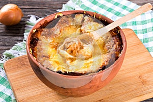 French onion gratin soup in a clay pot, authentic recipe, wooden spoon on a cutting board on an old rustic table, close-up