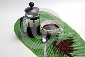 French press coffee with cup of coffee spoon and coarse coffee powder on banana leaf white background